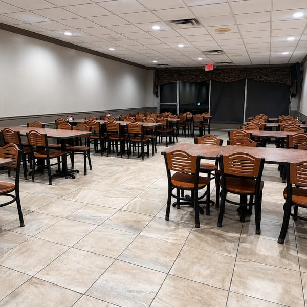 Empty restaurant dining room with many tables and chairs, light-colored tiled floor, and dark window coverings.