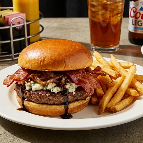 A bacon and blue cheese burger with fries, a pretzel, and a beer on a table.